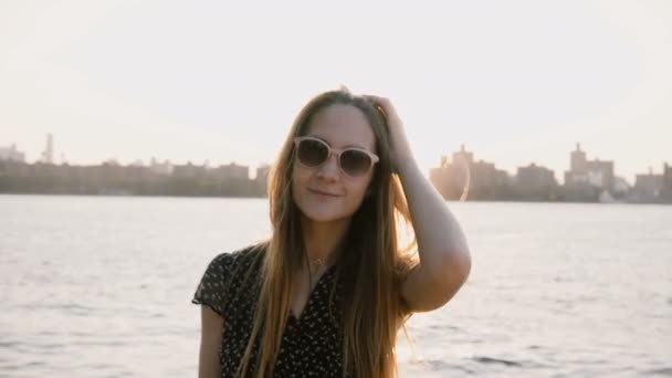 Feliz chica caucásica casual en gafas de sol de moda posando en la cámara, horizonte de la puesta del sol de Nueva York y el río en el fondo 4K . — Vídeos de Stock