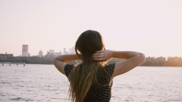 Achteraanzicht van meisje in zonnebril staan op zonsondergang strand, haren waait in de wind en de armen aan de orde gesteld in de lucht 4k. — Stockvideo