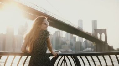 Kameraya 4 k seyir nehir setin çit Brooklyn köprüsünde eğilerek saç uçan ile güzel kız arkadan görünüm.
