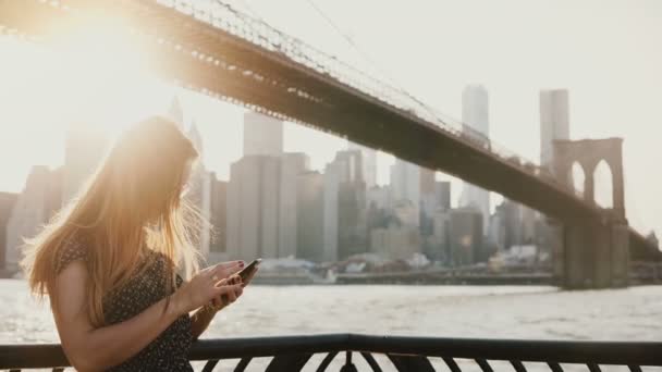 Beautiful girl with long hair in sunglasses using smartphone app at sunset river quay near Brooklyn Bridge New York 4K. — Stock Video