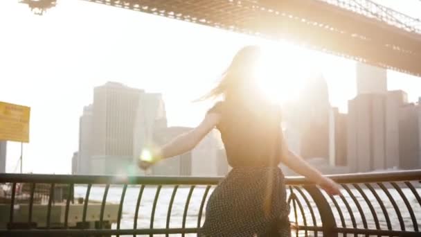 Hermosa chica viajera emocionada girando en el terraplén del río en el puente de Brooklyn con los brazos levantados en el aire en cámara lenta . — Vídeo de stock