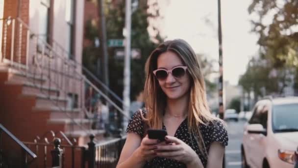 Menina europeia feliz com o cabelo longo sorrindo, andando ao longo de grandes edifícios de tijolos vermelhos velhos, usando aplicativo smartphone câmera lenta . — Vídeo de Stock