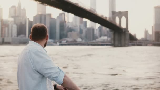 Vreedzame Kaukasische jongeman komt en staat op de dijk van de rivier in de buurt van New York City landschap, genieten van het uitzicht 4k. — Stockvideo