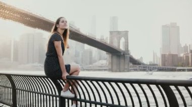 Güzel beyaz genç bir kadın nehir setin çit, Brooklyn Köprüsü'nün New York, 4 k tarafından geçen jetski oturan.