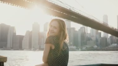 Poz, kamera günbatımı Brooklyn Köprüsü'nün Nehri Panoraması, Nyc 4 k yakınındaki gülümseyen mutlu oynak Avrupa Kız portresi. Casual genç heyecanlı kadın mükemmel yaz tatili zevk romantik ruh hali.