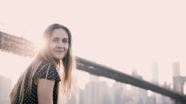 Atractiva chica caucásica mirando a la cámara sonriendo, sentada en la valla de terraplén Nueva York Brooklyn Bridge sunset view . — Vídeo de stock