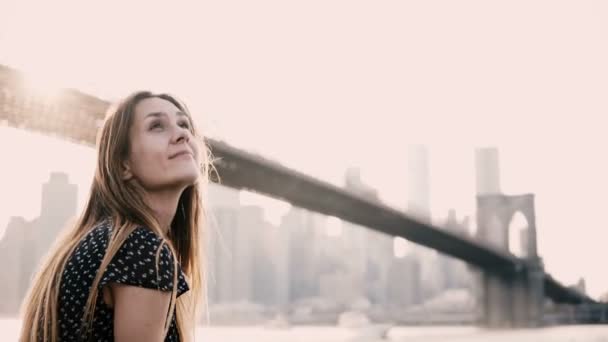 Heureuse jeune femme caucasienne souriante assise sur une clôture de remblai de rivière, profitant du panorama du pont de Brooklyn à New York 4K . — Video