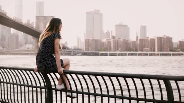 Hermosa chica europea sentada en la valla de terraplén del río, mirando hacia atrás a la cámara sonriendo cerca del puente de Brooklyn, NYC 4K . — Vídeos de Stock