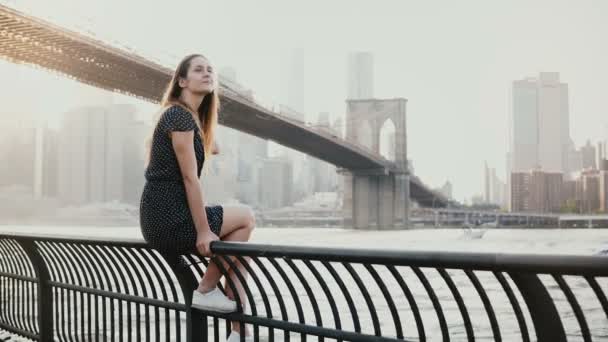 Kaukaska piękna młoda kobieta siedzi na płot nabrzeże rzeki w Brooklyn Bridge New York, jetski, przechodzącej przez 4k. — Wideo stockowe