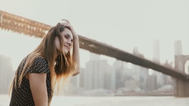 幸せの笑みを浮かべて欧州若い女性笑みを浮かべて、驚くべきニューヨークのブルックリン橋川 4 k 近くの夕景を楽しむ. — ストック動画