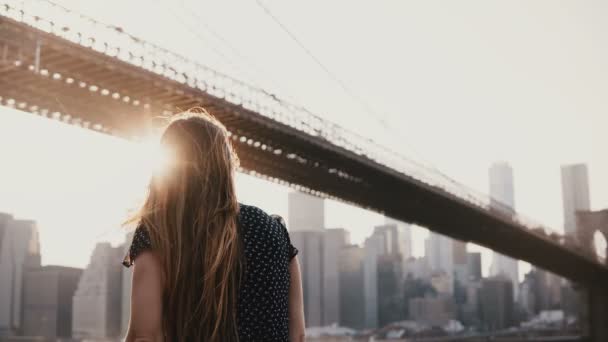 Vue arrière de la belle fille avec des cheveux soufflant dans le vent appuyé contre la clôture de remblai de la rivière près de Brooklyn Bridge 4K — Video