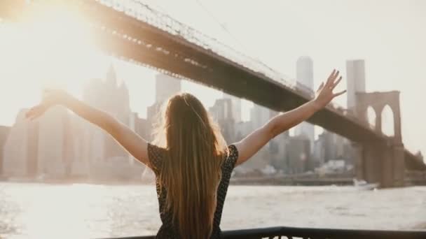 河川堤防、ニューヨークのブルックリン橋近く開いて腕に風立って吹くの髪の少女の背面図. — ストック動画