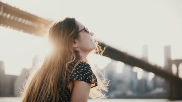 Щасливі усміхнений кавказьких молоду жінку в сонцезахисні окуляри, посміхаючись, насолоджуючись дивовижна Панорама річки захід сонця біля моста Брукліні 4 к. — стокове відео