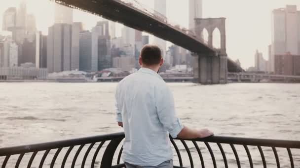 Réussi homme d'affaires heureux se tient près de Brooklyn Bridge remblai clôture panorama à New York, vue arrière 4K . — Video