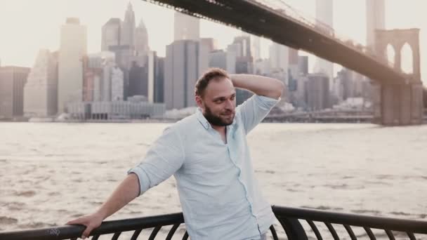 Homem caucasiano feliz fica perto da cerca de aterro do rio no cenário Brooklyn Bridge, refletindo e desfrutando da vista de Nova York — Vídeo de Stock