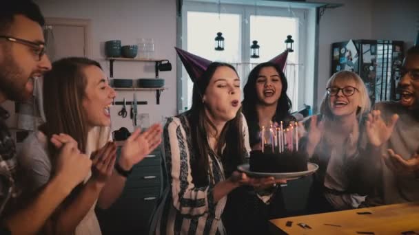Glückliches europäisches Mädchen hält Geburtstagstorte, bläst Kerzen an, feiert mit multiethnischen Freunden und Konfetti Zeitlupe. — Stockvideo