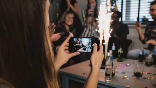 Chica tomando una foto de teléfono inteligente de la fiesta de cumpleaños con chispeantes fuegos artificiales y amigos multiétnicos aplaudiendo cámara lenta . — Vídeos de Stock