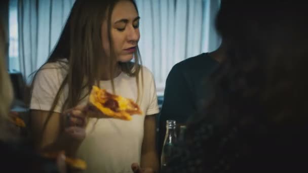 Giovane bella ragazza caucasica mangiare pizza con gli amici a un tavolo da bar della cucina in una festa casalinga divertente casa con bevande . — Video Stock