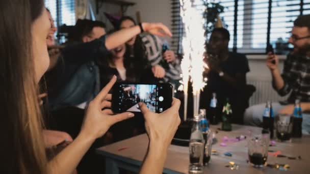 Las manos femeninas toman una foto de teléfono inteligente de la fiesta de cumpleaños multiétnica con velas de fuegos artificiales, amigos abrazando cámara lenta . — Vídeo de stock