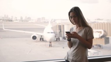Üzgün ve sinirli bakarak telefon ve akıllı seyretmek Havaalanı terminal penceresi duran genç Avrupa kadın alır.