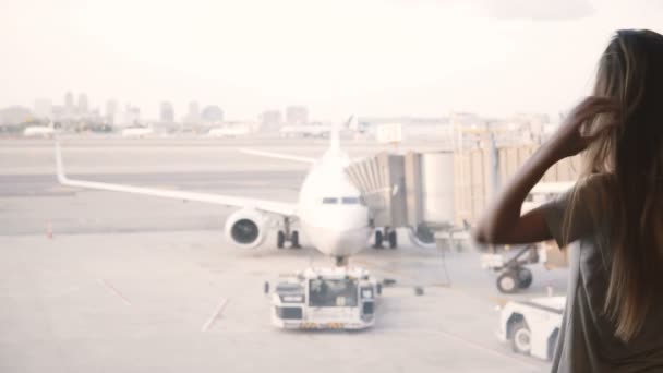 Hermosa chica caucásica con el pelo largo camina hasta la ventana terminal del aeropuerto y espera, avión en el fondo . — Vídeos de Stock