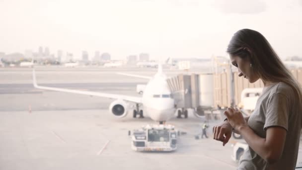 Jonge Kaukasische zakenvrouw permanent op de luchthaven terminal lounge venster met behulp van slimme horloge, vliegtuigen te kijken. — Stockvideo