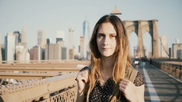 Jonge positieve vrouwelijke blanke toerist met rugzak kijken camera, glimlachend op zonnige Brooklyn Bridge, New York 4k. — Stockvideo