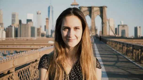 Mladý Evropský krásné cestovatel dívka dotýká vlasů, při pohledu na fotoaparát a usmívá se na Brooklynský most v New Yorku 4k — Stock video