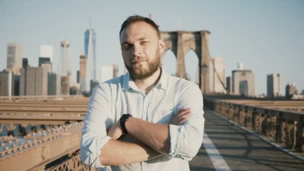 Молодий позитивні чоловічого кавказьких бізнесмен з обіймами перетнула дивлячись на камеру, посміхаючись на Бруклінський міст, Нью-Йорк 4 к. — стокове відео