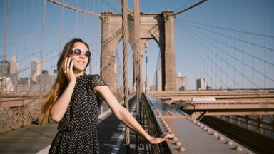 Telefonda, Brooklyn köprüsünde 4 k uzun saç esen rüzgar retro güneş gözlüğü görüşmelerde mutlu Avrupa işkadını.