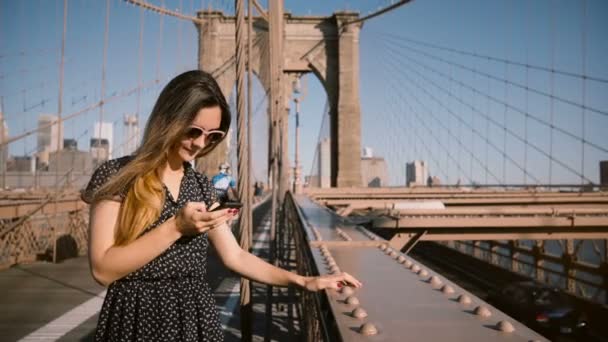 Hermosa mujer blogger europea sonriente con gafas de sol elegantes usando la aplicación para teléfonos inteligentes en Brooklyn Bridge, alejándose 4K . — Vídeo de stock