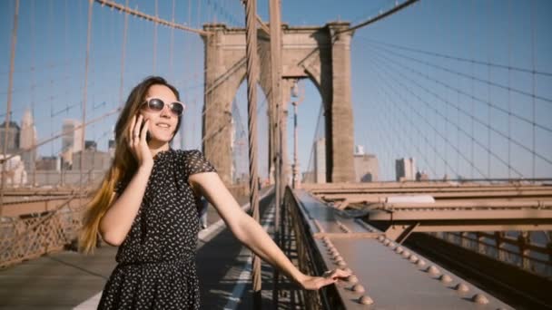 Happy Evropské podnikatelka v retro sluneční brýle mluví o telefonu, vítr vane v dlouhé vlasy na Brooklyn Bridge 4k. — Stock video