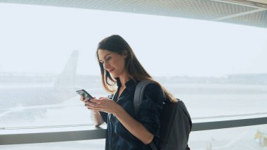 Havaalanı pencere Smartphone kullanan genç kız. Sırt çantası ile mutlu Avrupa kadın terminalde mobil uygulaması kullanır. 4k.