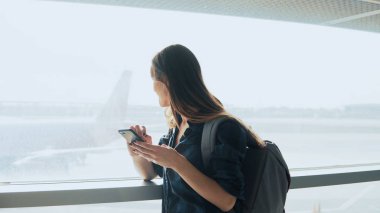 Havaalanı pencere Smartphone kullanan genç kız. Sırt çantası ile mutlu Avrupa kadın terminalde mobil uygulaması kullanır. 4k.