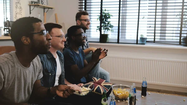 Mixed race male supporters watch sports on TV 4K. Fans celebrate goal victory holding British flag cushion. Slow motion.