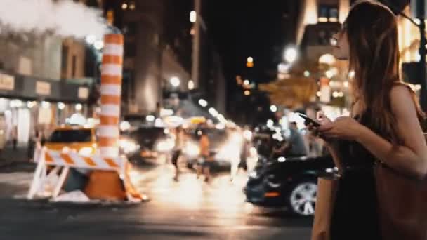 Joven hermosa mujer cruzando la carretera de tráfico en el centro de Nueva York, Estados Unidos por la noche y utilizando un teléfono inteligente . — Vídeos de Stock