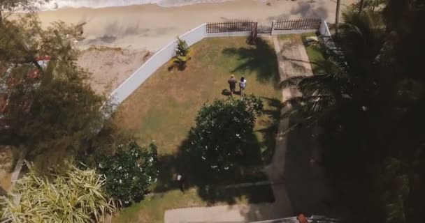 Drone voando para a frente sobre pequena casa de resort tropical idílico, casal caminhando lentamente para a costa do mar lavado por ondas . — Vídeo de Stock