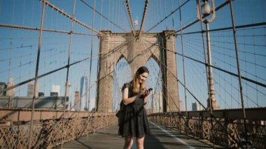 Brooklyn Köprüsü, New York Smartphone kullanarak genç mutlu beyaz bir kadın. Kadın işkadını alışveriş app 4 k kullanır.