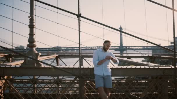 Glad kaukasiska affärsman står vid Brooklyn Bridge, New York skriver på smart watch armband, sedan på smartphone 4k. — Stockvideo