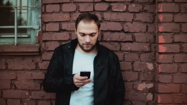 Homme caucasien occasionnel utilisant un smartphone à l'extérieur. Beau barbu jeune homme d'affaires textos. Mur en brique rouge 4K . — Video