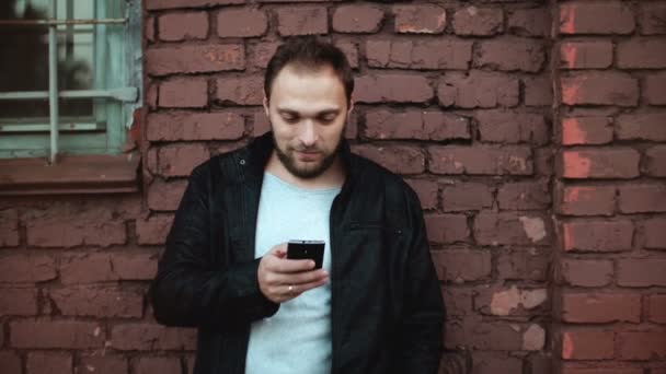 Zrelaksowany człowiek Europy za pomocą aplikacji na smartfony poza. Przystojny Brodaty mężczyzna biznesmen mobilne biuro. Czerwony mur z cegły 4k. — Wideo stockowe