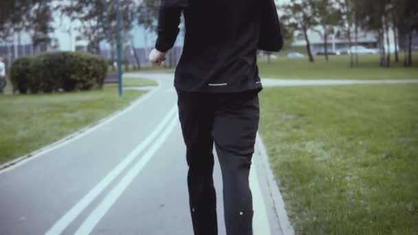 Un homme courant le long d'une route de parc. Au ralenti. Vue de derrière. Inclinez-vous. Un homme d'affaires prospère faisant du jogging dans une allée verte . — Video