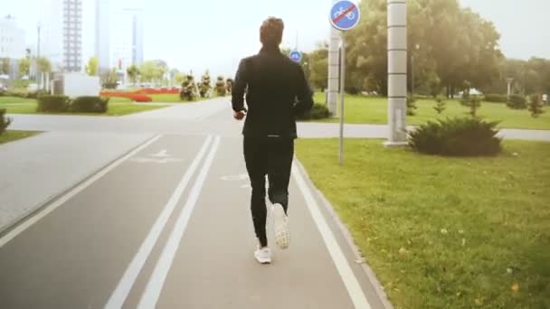 4K Homme courant le long d'une route de parc de la ville. Vue de derrière. Fitness runner en vêtements de sport noirs, baskets blanches. Mode de vie sain . — Video