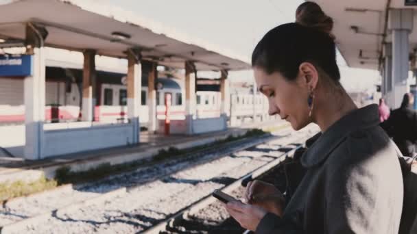 Young European businesswoman typing messages on smartphone at railway station, stressed and late, train passing by. — Stock Video