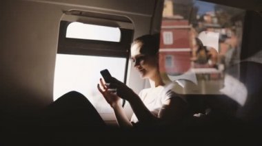 Gülümseyerek, hareketli modern tren pencere koltukta oturan smartphone app kullanarak mutlu Avrupa kız inanılmaz yansıma çekim.