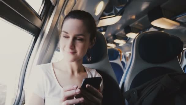 Vista de cerca de la hermosa chica caucásica sonriente feliz usando el teléfono inteligente, mirando por la ventana del tren y alejándose . — Vídeo de stock