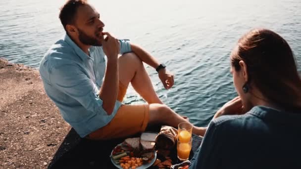 Junges glückliches Paar, das am Ufer eines Meeres sitzt und isst. Mann und Frau picknicken morgens am Wasser. — Stockvideo