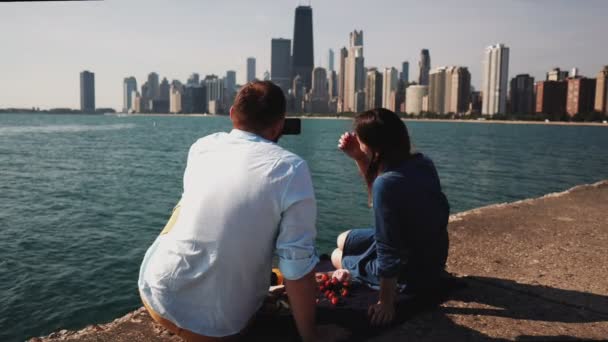 Chicago, Amerika üzerinde Michigan Gölü kıyısına piknik genç çiftin arkadan görünüm. Adam fotoğraf Smartphone'da alır. — Stok video