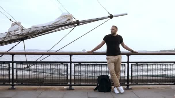 Schöner junger Mann, der auf der Seebrücke in der Nähe des Zauns nahe dem Bugspriet des Schiffes steht und sich umsieht. — Stockvideo