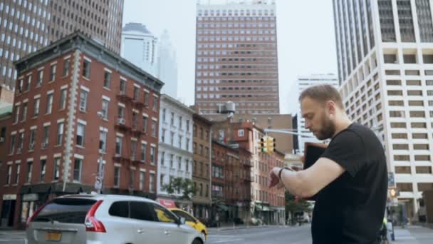 Pemuda tampan berdiri di dekat jalan dan menggunakan jam tangan pintar di distrik keuangan di New York. Gerakan lambat . — Stok Video